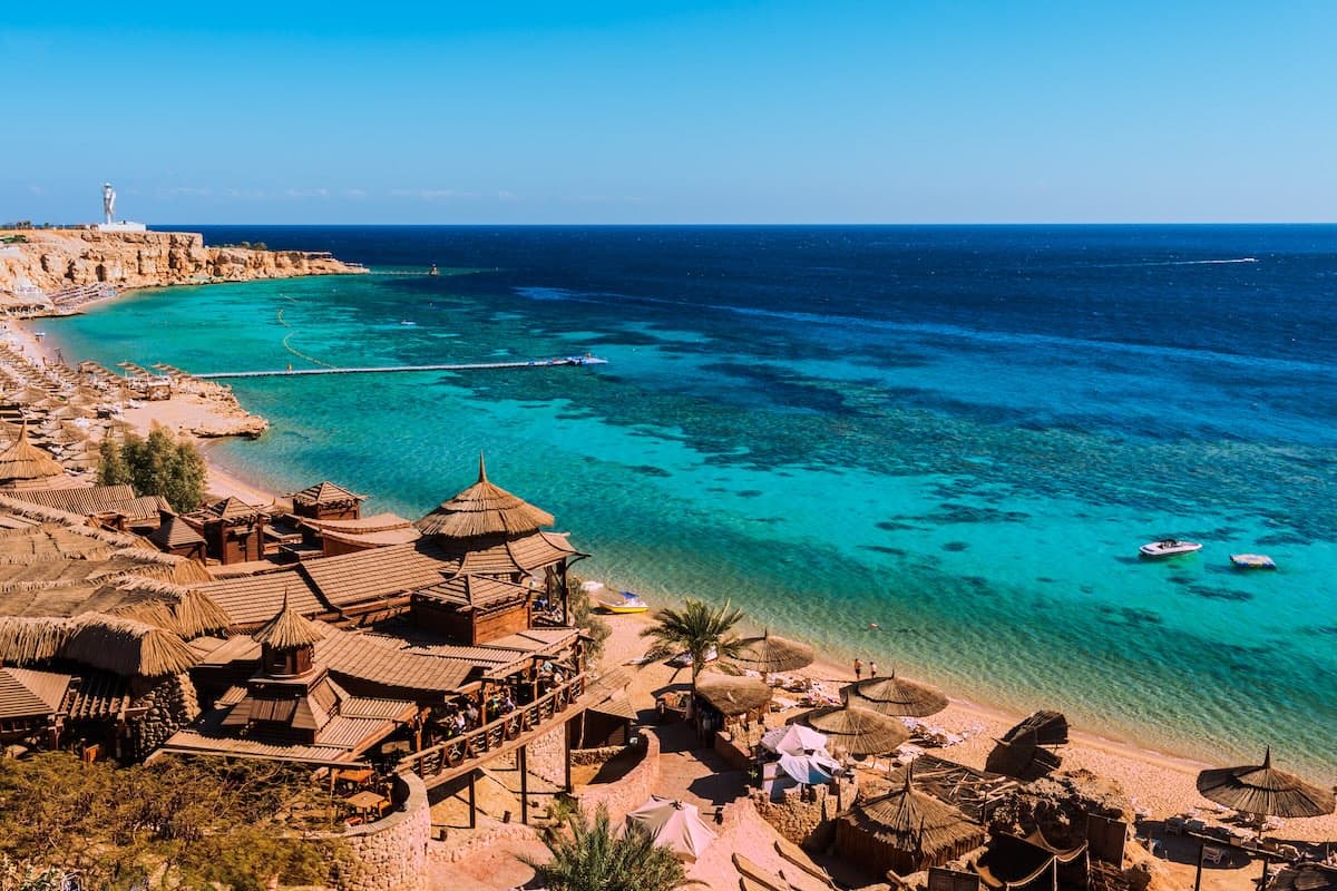 Vista aérea de una bahía en Sharm el-Sheikh junto al Mar Rojo — restaurantes de playa sobre pilotes, aguas turquesas con arrecifes de coral visibles, embarcaderos y lagunas.